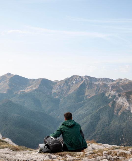 a photo of the back of seated person wearing a jacket with a backpack, gear and a sleeping mat setting next to him, pondering the awesome view of rugged mountains ahead of him

